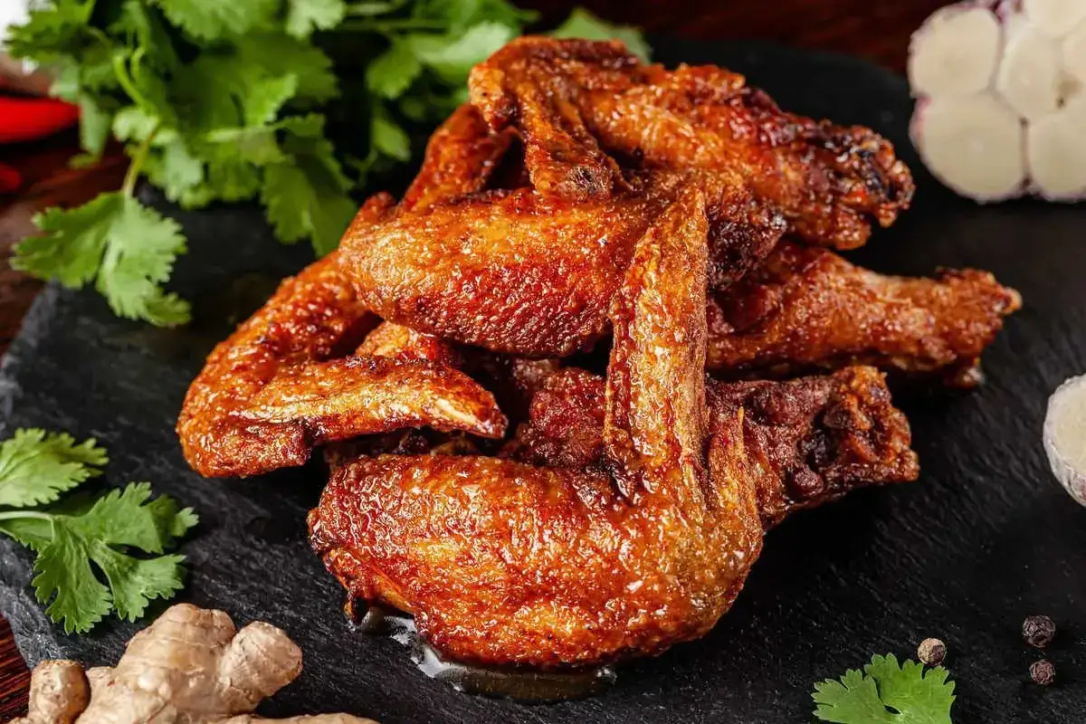 Alette di pollo dorate e croccanti, servite su una pietra scura con zenzero, aglio e coriandolo fresco.