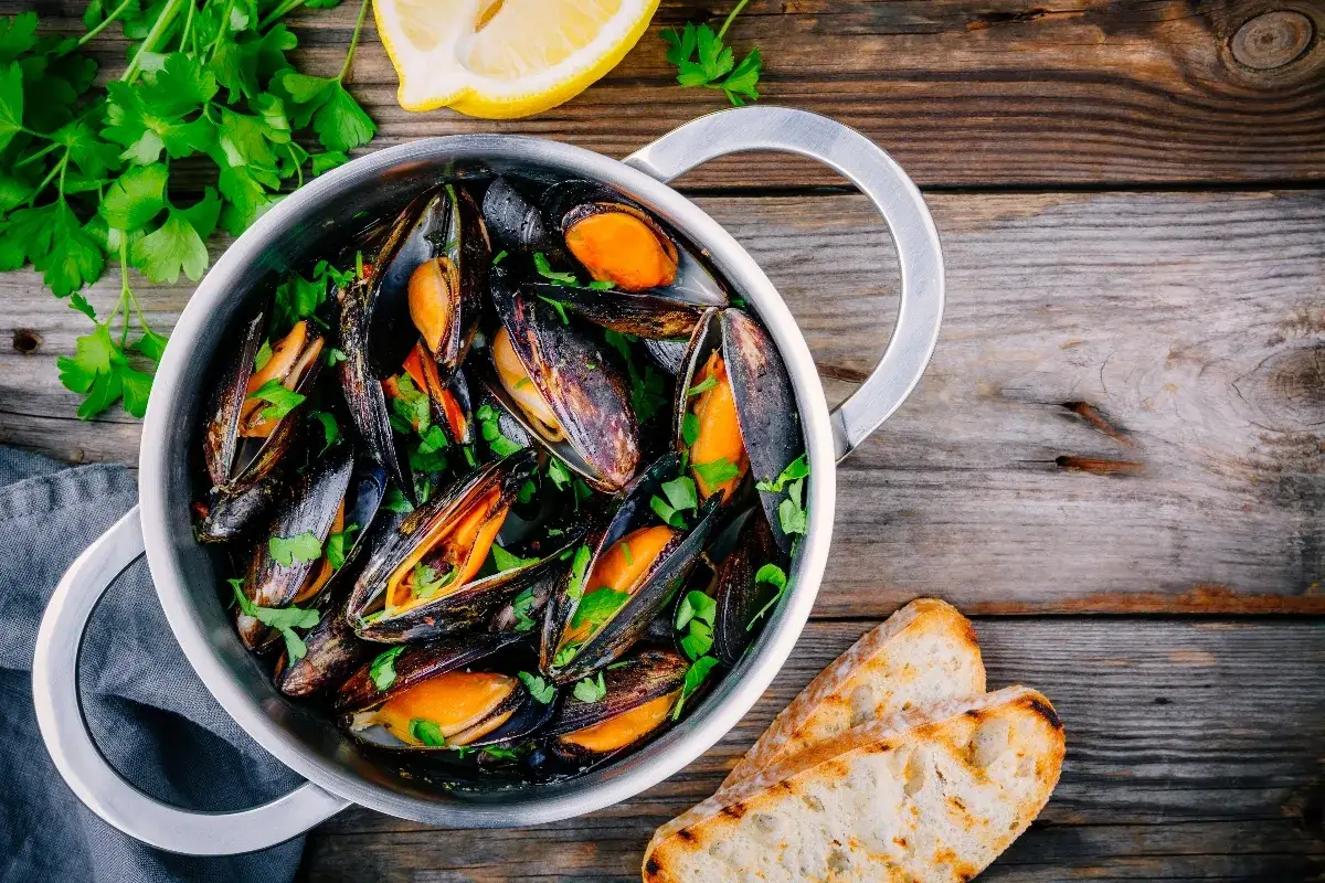 Un pentolino pieno di cozze alla marinara, guarnite con prezzemolo fresco e accompagnate da fette di pane tostato e spicchi di limone.