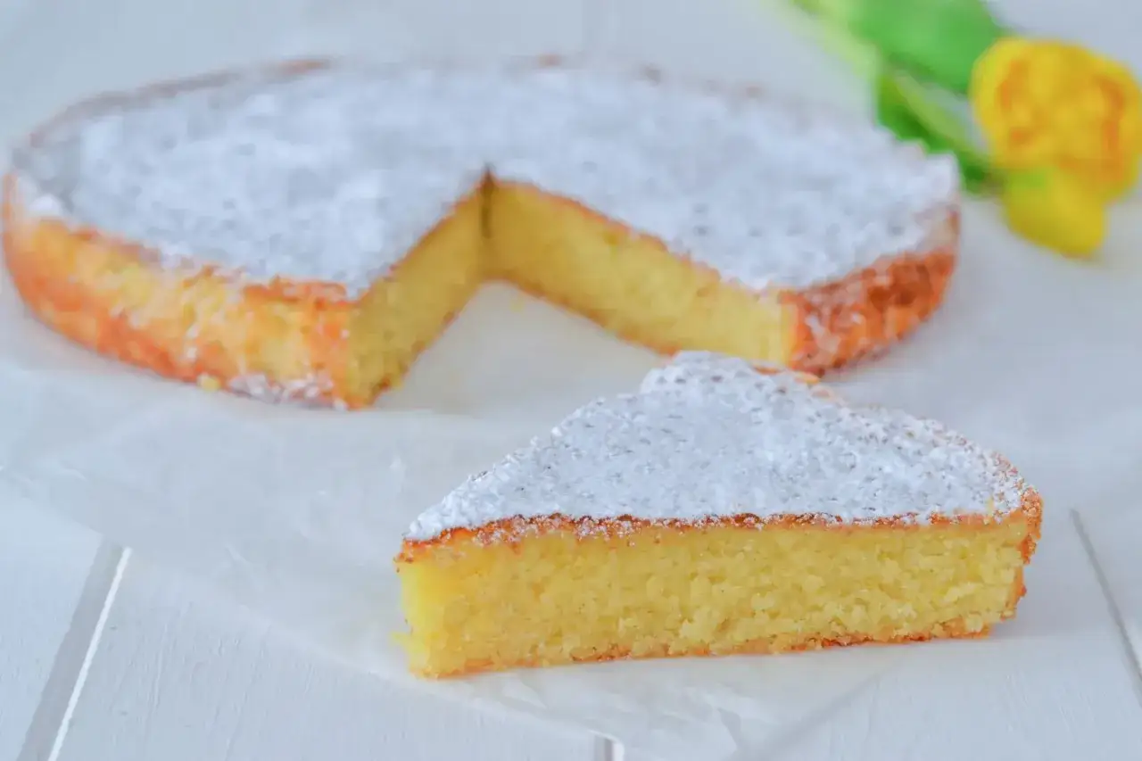 Una fetta di torta caprese al limone, soffice e cosparsa di zucchero a velo, accanto alla torta intera.