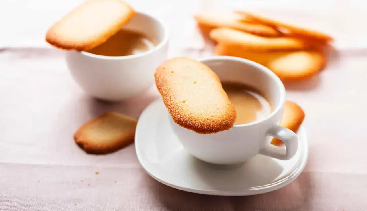 Deliziose lingue di gatto dorate appoggiate su tazze di caff&egrave; fumante, un invito alla dolcezza.