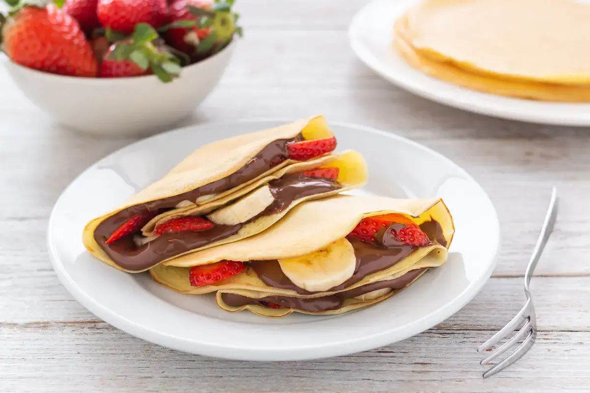 Crepes alla Nutella con fragole e banane, un dolce invitante su un piatto bianco, con una ciotola di fragole sullo sfondo.