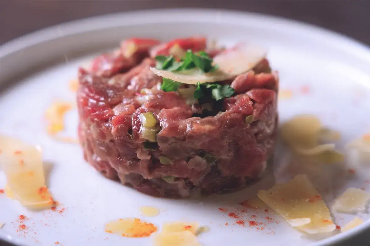 Deliziosa tartare di manzo finemente tritata, guarnita con erbe fresche e scaglie di parmigiano su un piatto bianco.