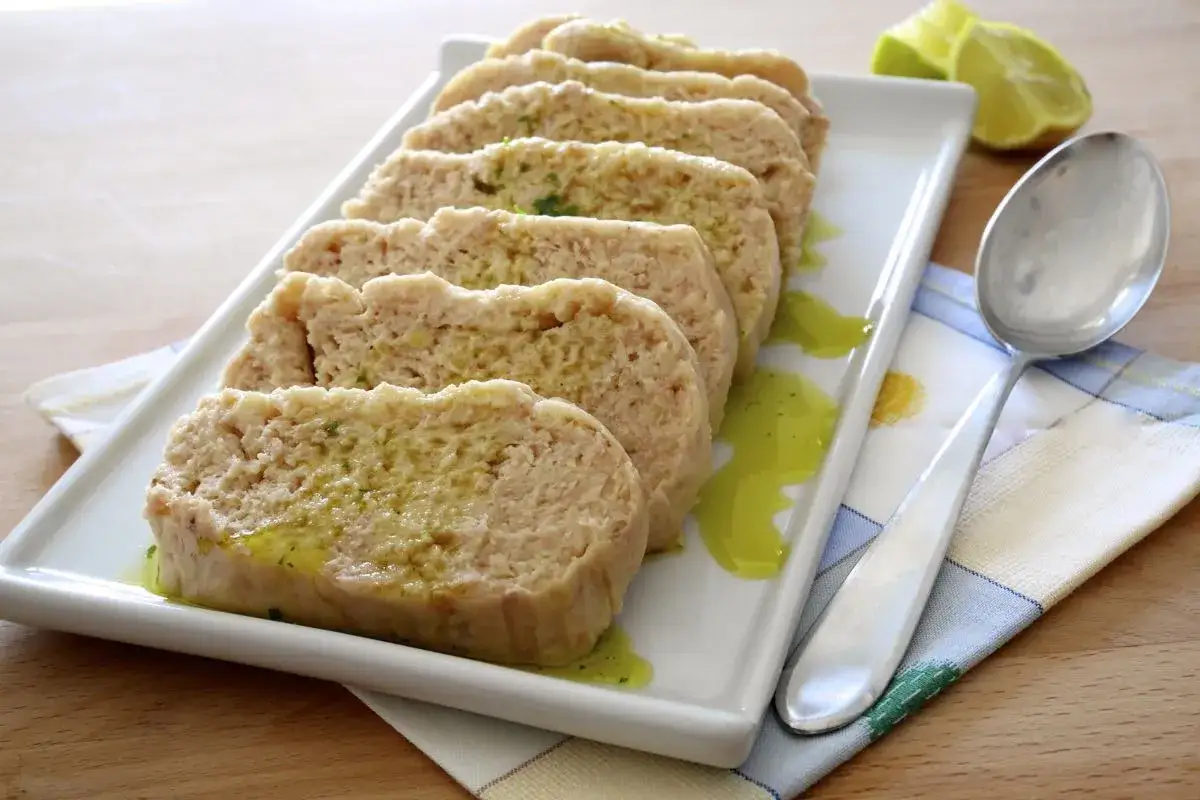 Fette di polpettone di tonno condite con olio e erbe, servite su un piatto bianco con cucchiaio e spicchi di lime.