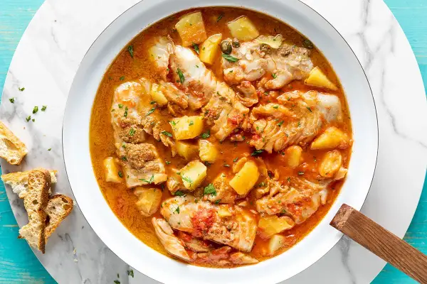 Un invitante merluzzo in umido con patate, pomodoro e capperi, guarnito con prezzemolo fresco e accompagnato da fette di pane croccante.