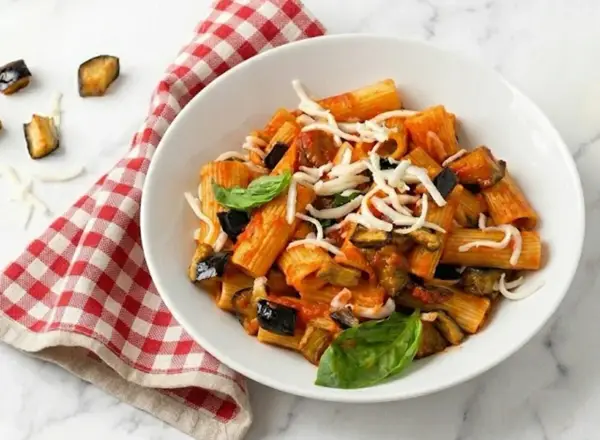 Un piatto di pasta alla norma fumante, con melanzane fritte, pomodoro e ricotta salata grattugiata, guarnito con basilico fresco.