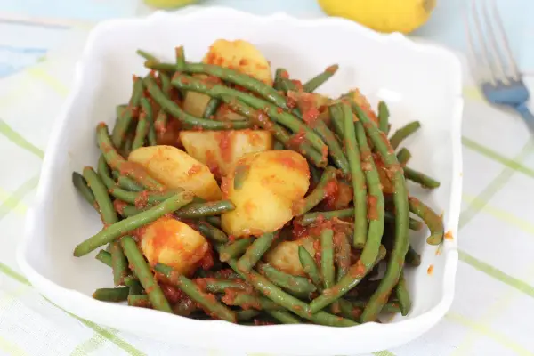 Un piatto di fagiolini in umido con patate, conditi con un sugo rosso saporito, servito in una ciotola bianca.