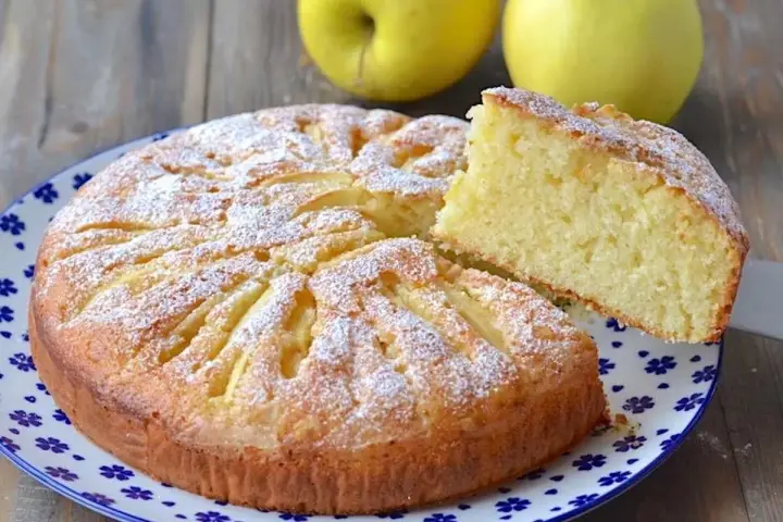 Deliziosa torta di mele con yogurt, cosparsa di zucchero a velo, con una fetta sollevata per mostrare la morbidezza.