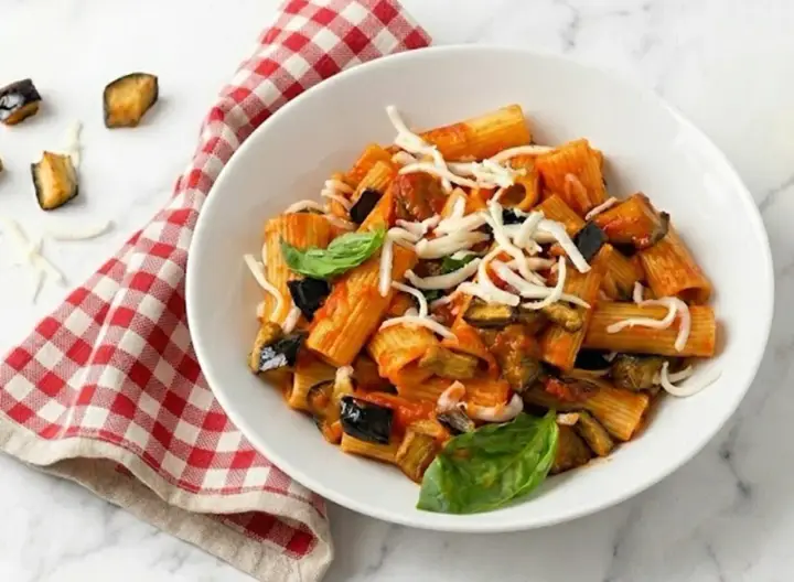 Un piatto di pasta alla norma fumante, con melanzane fritte, pomodoro e ricotta salata grattugiata, guarnito con basilico fresco.