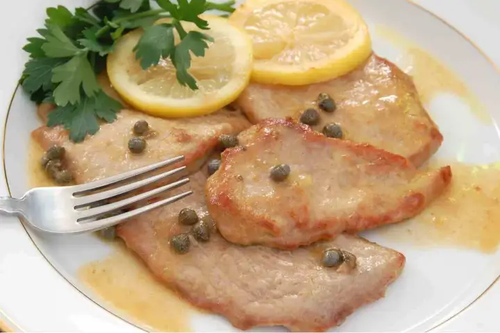 Scaloppine al limone succulente, guarnite con capperi, prezzemolo e fette di limone, pronte per essere gustate con una forchetta.