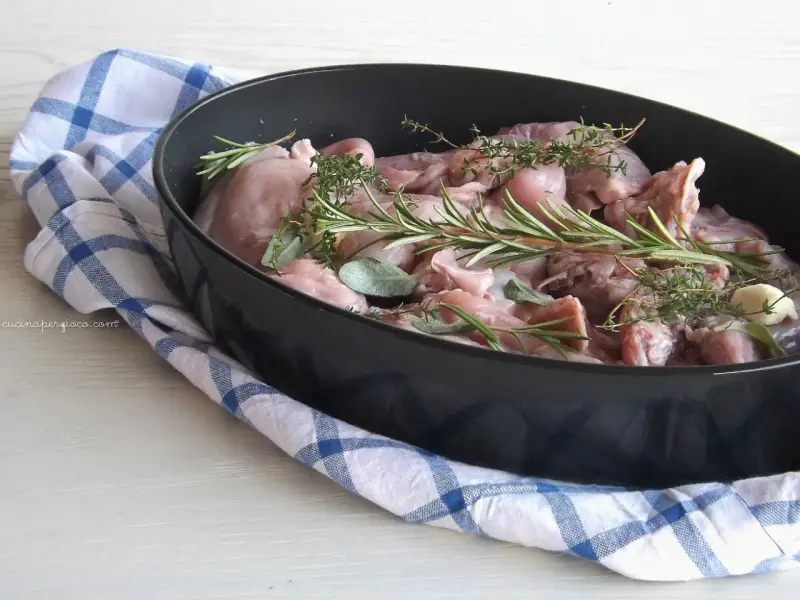 Pezzi di coniglio crudo in una teglia, pronti per essere cucinati al forno con rosmarino, salvia e aglio.