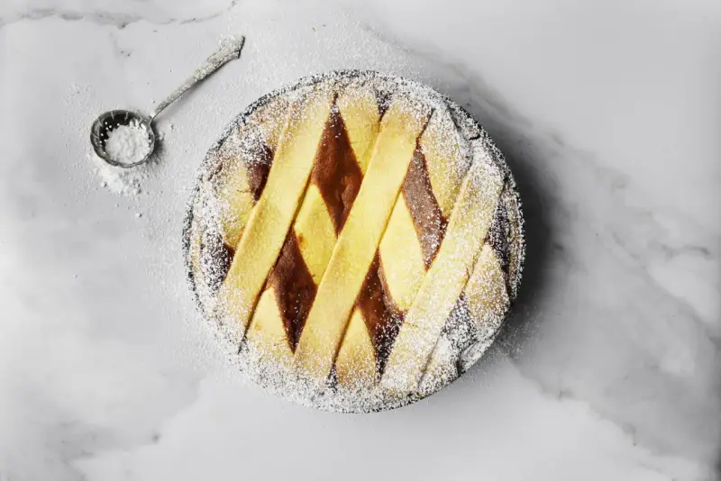 Pastiera napoletana pronta, cosparsa di zucchero a velo. Un cucchiaino d'argento è pronto per decorare, mostrando come si fa la pastiera.