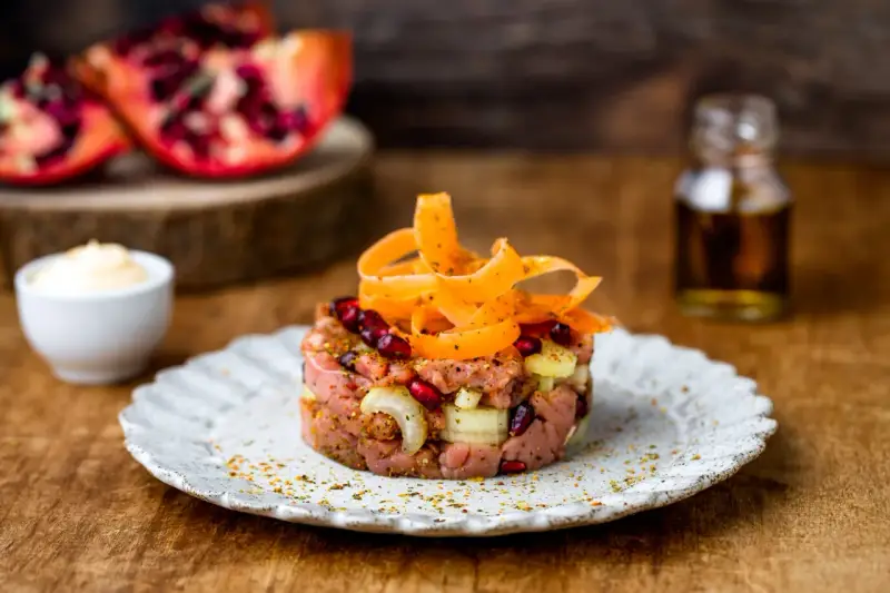 Tartare di manzo con melograno e carote, servita su un piatto rustico. Sullo sfondo, spicchi di melograno e una bottiglietta di condimento.