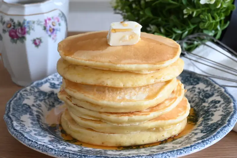 Pila di soffici pancake con farina di avena, guarniti con burro e sciroppo d'acero, su un piatto decorato.