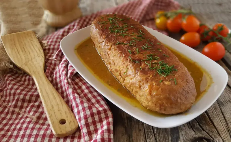 Polpettone in padella dorato e succulento, guarnito con erba cipollina, pronto per essere servito. Accanto, una spatola in legno e pomodorini freschi.