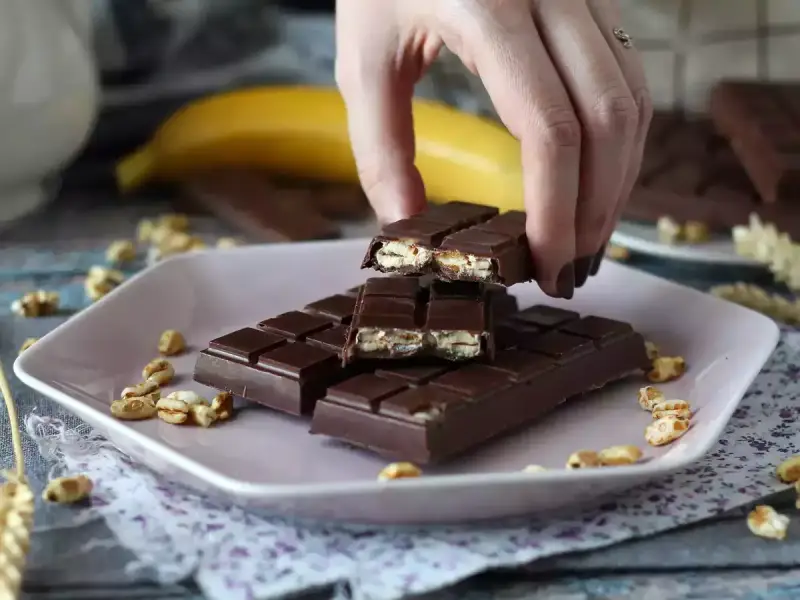 A hand breaks a chocolate bar filled with puffed rice and nuts on a pink plate.